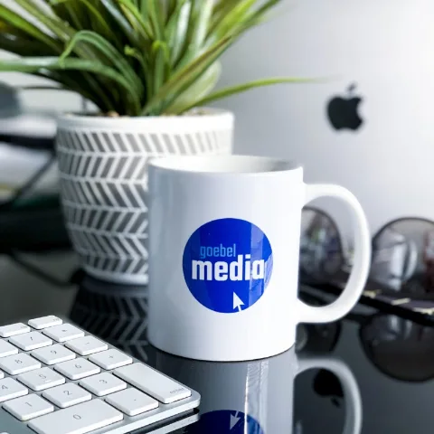 A Goebel Media mug on a studio desk beside a keyboard and a plant