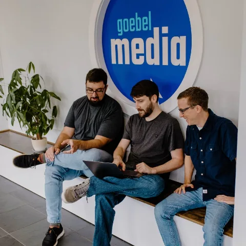 Three team members reviewing work on a laptop under the Goebel Media wall logo